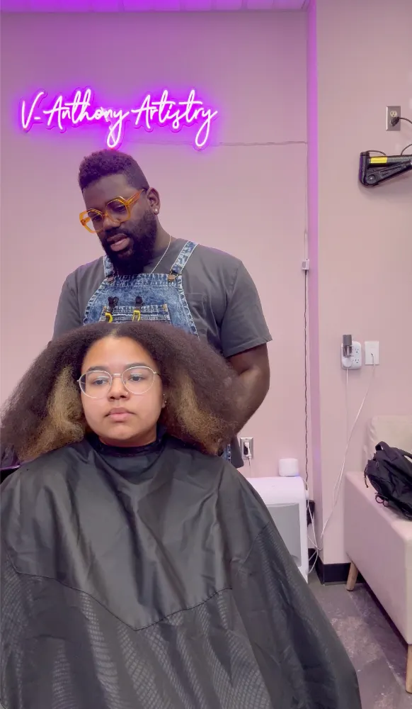 Victor in denim overalls standing behind a seated guest with long, two-tone natural hair, talking through the plan before anything starts